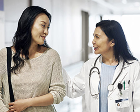 Smiling mature doctor talking to woman in hospital. Female patient visiting healthcare worker for routine checkup. They are standing in corridor.