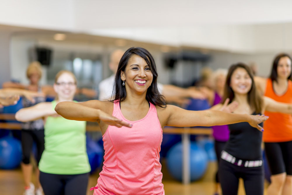 A woman is leading out in a fitness dance class at her indoor studio. She is teaching a multi-ethnic class that is wearing fitness clothing and dancing to the music.