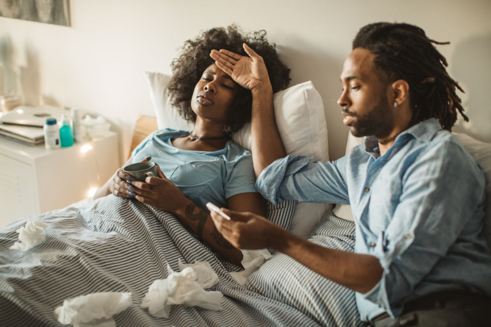 Mujer afroamericana joven sintiéndose enferma en casa. Acostada en la cama, bebiendo té y medicinas. Su novio o esposo la cuida. De día.