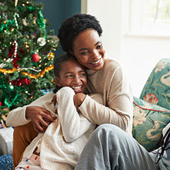 Smiling young woman embracing girl while sitting on sofa at home during Christmas festival