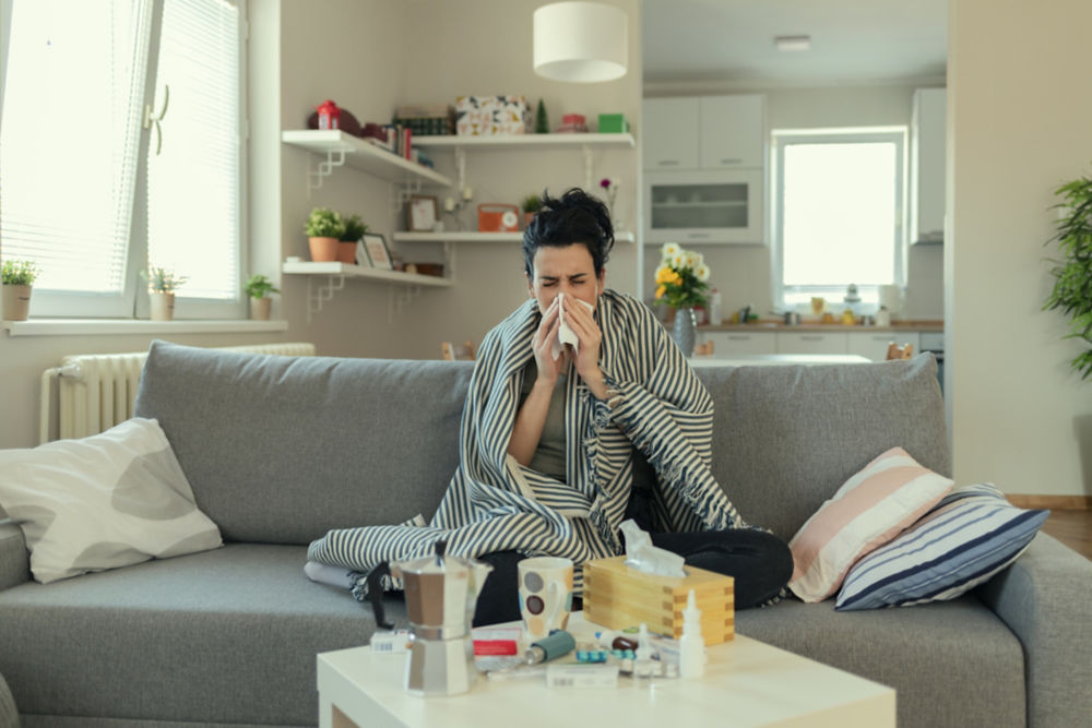 Mujer enferma cubierta con una manta acostada en la cama con fiebre alta y flu, descansando en la sala de estar. Ella está agotada y sufre del flu. Mujer enferma con secreción nasal acostada en la cama. Niña sufriendo de frío acostada en la cama con un pañuelo sonándose la nariz mientras está sentada en el sofá