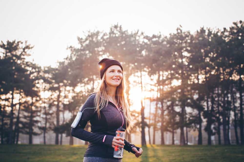 Mujer joven trotando afuera en un bosque de otoño soleado. Se toma un descanso y bebe agua. El sol brilla en el fondo.