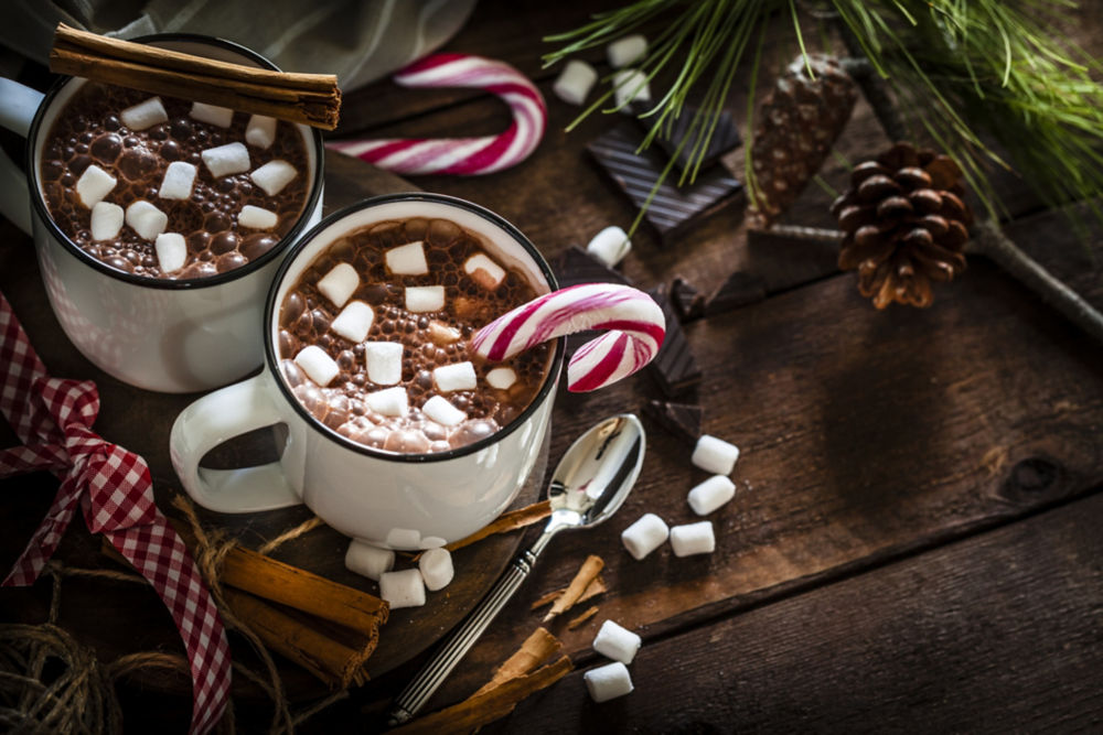 Vista de imagen de ángulo alto de dos tazas de chocolate caliente caseras con malvaviscos sobre una mesa navideña de madera rústica. Un bastón de caramelo está dentro de una de las tazas y otro colocado directamente sobre la mesa. Una decoración navideña completa la composición. Foto de estudio DSRL de bajo perfil tomada con cámaras Canon EOS 5D Mk II y Canon EF 100 mm f/2.8L Macro IS USM