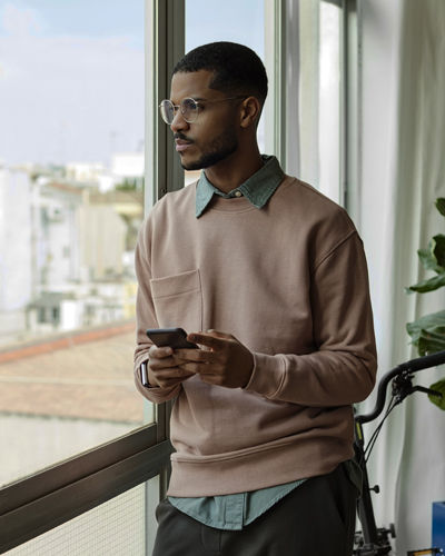 Handsome young man looking through window. Thoughtful male is with mobile phone in living room. He is at home office.