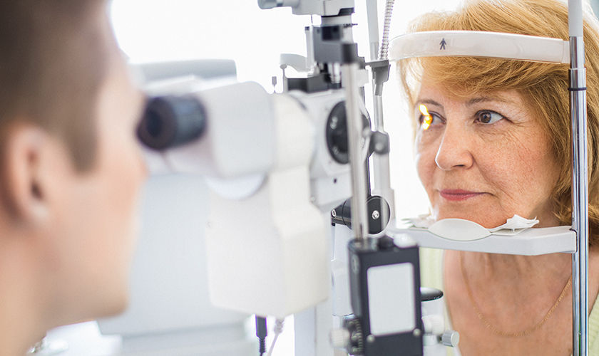 Una mujer mayor caucásica a la que le están realizando un examen de la vista en la óptica. La cabeza está apoyada en un aparato foróptero mientras un médico varón de mediana edad le examina la retina. La mujer tiene el cabello castaño claro de longitud media y lleva una blusa de color verde claro.