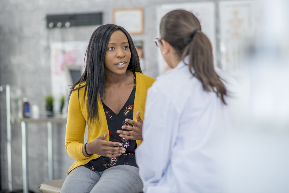 A woman and her doctor are indoors in a medical clinic. The woman is sitting and describing her symptoms to the doctor.