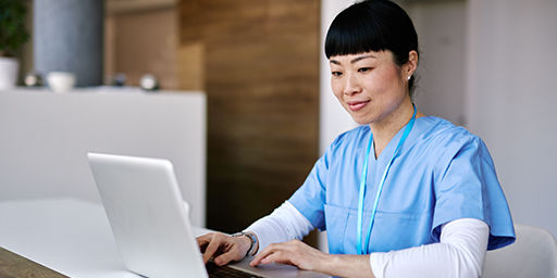 Smiling mid adult asian female nurse in blue scrubs is sitting at a white desk typing on a laptop, with a clipboard and documents in the foreground, in a brightly lit hospital room