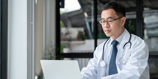 A professional senior male doctor is working on his laptop computer in his office at the clinic, reading medical cases online or responding to emails.