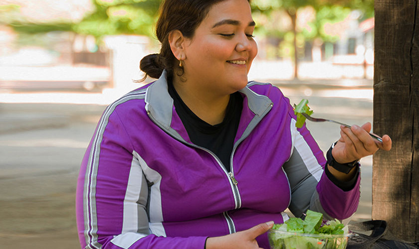 Mujer feliz con sobrepeso sonriendo mientras come una ensalada verde saludable antes de comenzar sus ejercicios de cardio al aire libre