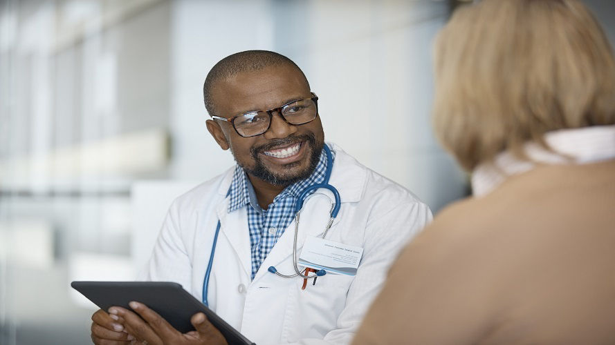 Médico sonriente sosteniendo una tableta digital mientras observa a una anciana. El paciente está visitando a un trabajador de cuidado de salud en el hospital. Lleva una bata de laboratorio y anteojos.