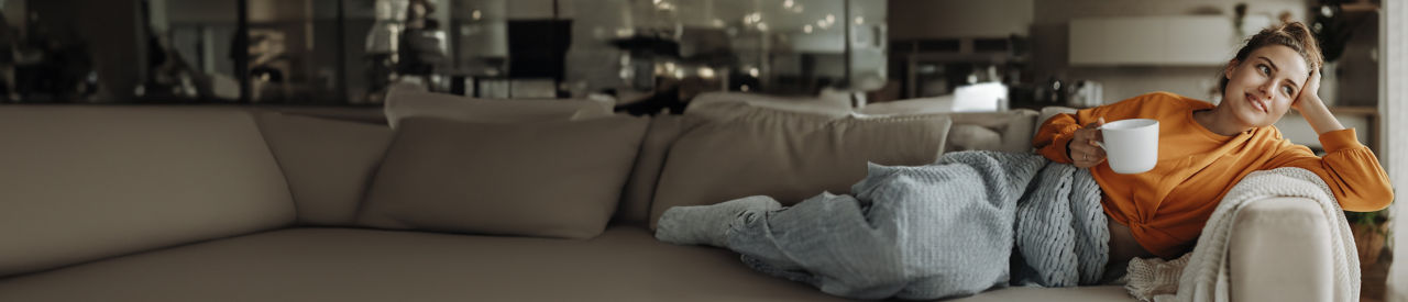 woman sitting on couch with blanket and drinking coffee while looking out the window
