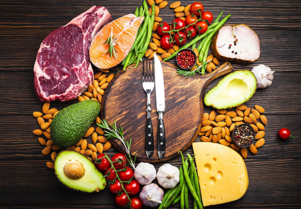 Knife and fork over wooden cutting board and ketogenic low carbs ingredients for healthy eating concept and weight loss, top view. Keto foods: meat, fish, avocado, cheese, vegetables, nuts