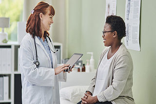 Smiling mature doctor talking to woman in hospital. Female patient visiting healthcare worker for routine checkup. They are standing in corridor.