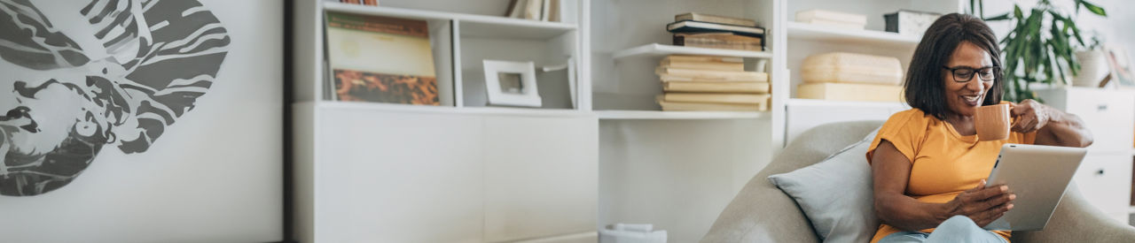 Senior black woman drinking from a mug and using digital tablet while sitting in living room at home.