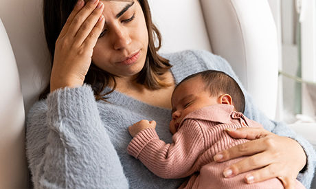 Mother sitting on couch in nursery feeling postpartum depression with baby in her arms.