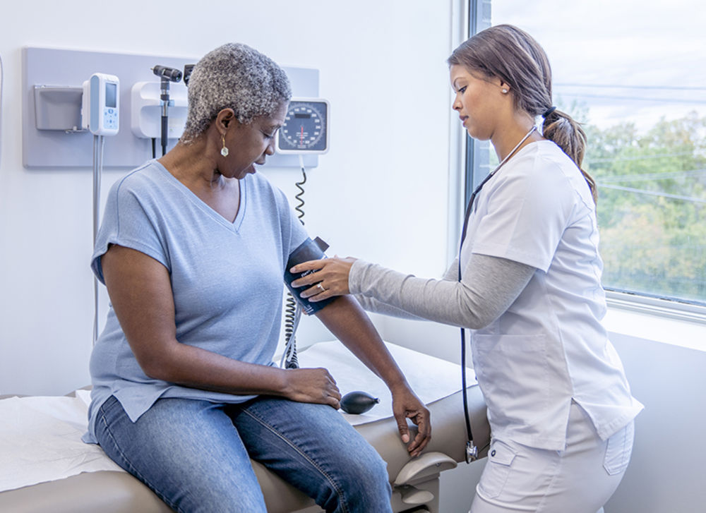 Una paciente mayor de ascendencia africana se sienta en una mesa de examen mientras su médica le toma la presión arterial.  Está vestida de manera informal y mira abajo hacia el brazo mientras la médica sostiene el manguito de presión arterial.