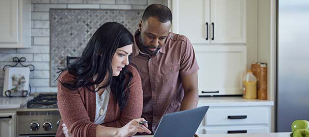 two people sit at kitchen counter with breakfast working with pen, paper and laptop.