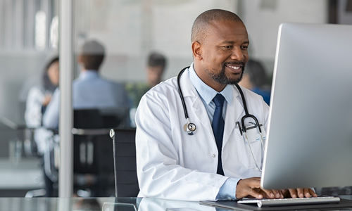 Practitioner typing on computer in modern clinic. Doctor at work in office using computer with copy space. African happy specialist wearing white coat and sitting at desk while mediacal team having meeting in background.