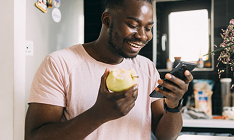 Un hombre sonriente comiendo un refrigerio saludable mientras le envía un mensaje de texto a su asesor de salud en su teléfono celular