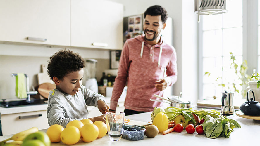 Kid and his dad are in the kitchen. They are making healthy food with vegetables. 